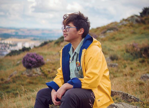 A photo of Nemo Martin. They are an Asian person with short brown hair and glasses. They are sitting on a rock on a grassy mountain face. They are wearing a bright yellow raincoat and looking into the distance.
